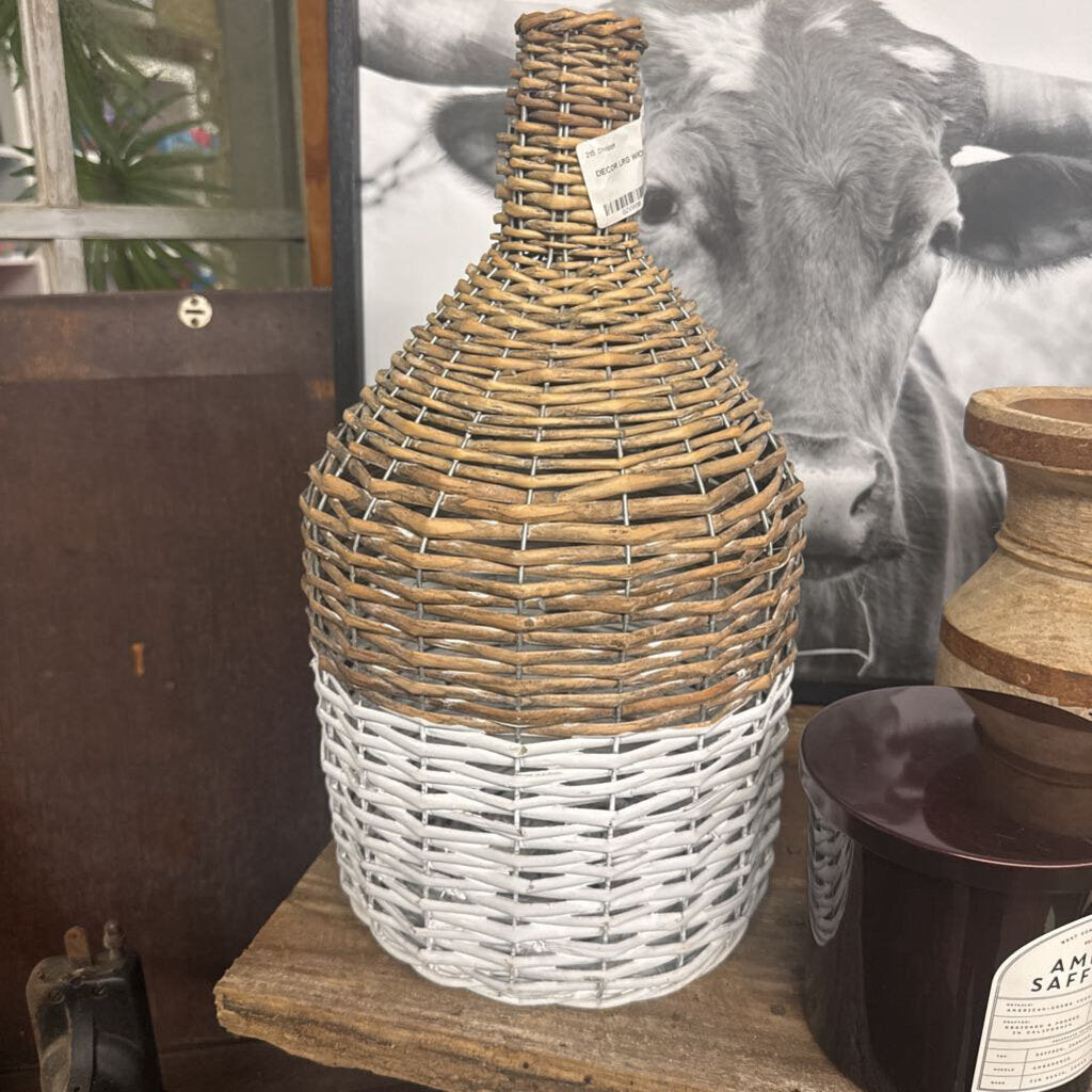 A decorative woven bottle-shaped basket with a natural tan top half and a white-painted bottom half, displayed on a wooden surface in an indoor setting.