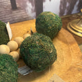 Three green moss-covered decorative balls displayed on a wooden surface, with small light wood beads and a tag visible nearby.
