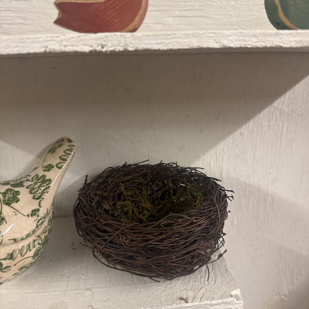 A small decorative twig nest with green moss sits on a white painted shelf next to a ceramic bird figurine.