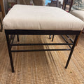 An upholstered stool with a cream bouclé cushion and a black metal frame, shown on a woven jute rug in a home setting.