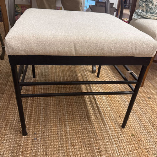 An upholstered stool with a cream bouclé cushion and a black metal frame, shown on a woven jute rug in a home setting.