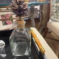 A clear glass decanter bottle with a cork stopper and a decorative pinecone insert, placed on a black tray next to a bamboo-handled tool in a home setting.