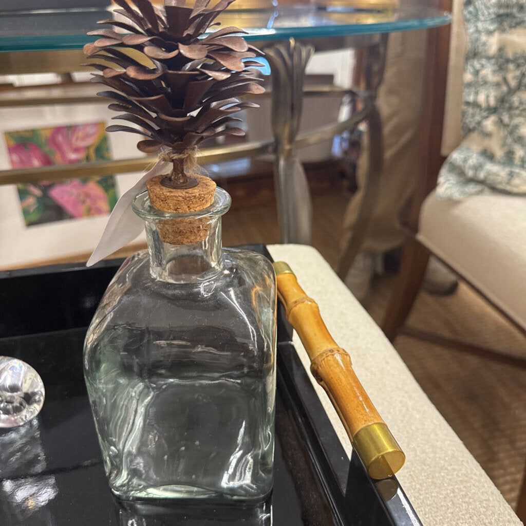 A clear glass decanter bottle with a cork stopper and a decorative pinecone insert, placed on a black tray next to a bamboo-handled tool in a home setting.