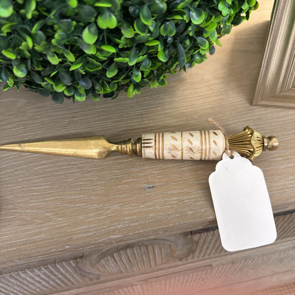 A brass and carved bone letter opener with a blank white tag attached, resting on a wooden surface next to a green boxwood top.