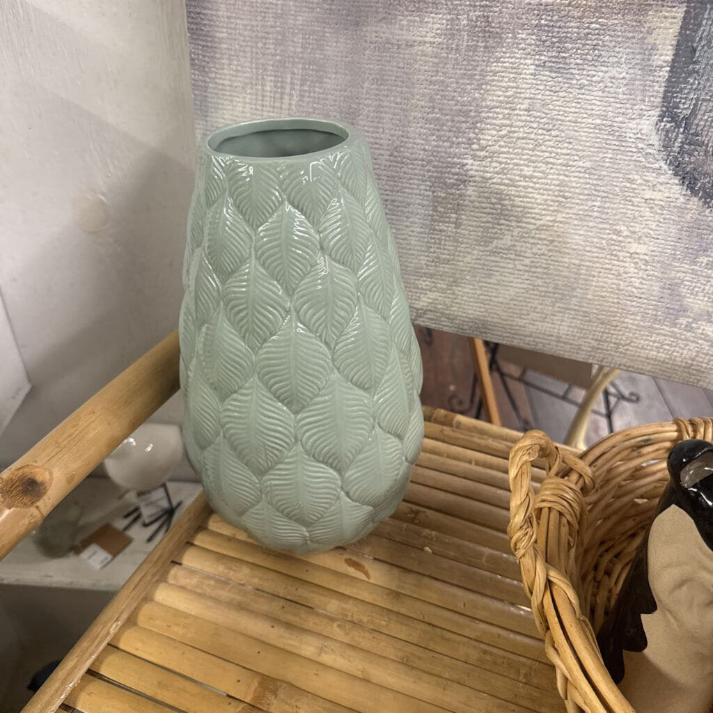A light sage green ceramic vase with a textured, quilted leaf pattern, displayed on a bamboo slatted tray next to a wicker basket.