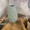 A light sage green ceramic vase with a textured, quilted leaf pattern, displayed on a bamboo slatted tray next to a wicker basket.