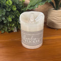 A flameless LED candle in a clear plastic wrap with a gray paper label reading 'flameless flicker' on a wooden table with green plants in the background.