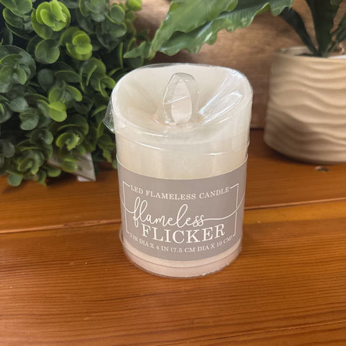 A flameless LED candle in a clear plastic wrap with a gray paper label reading 'flameless flicker' on a wooden table with green plants in the background.