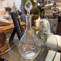 A clear glass decanter with a matching faceted glass stopper, decorated with etched scallop patterns on the bulbous body, displayed on a glass-topped table in a home decor store.