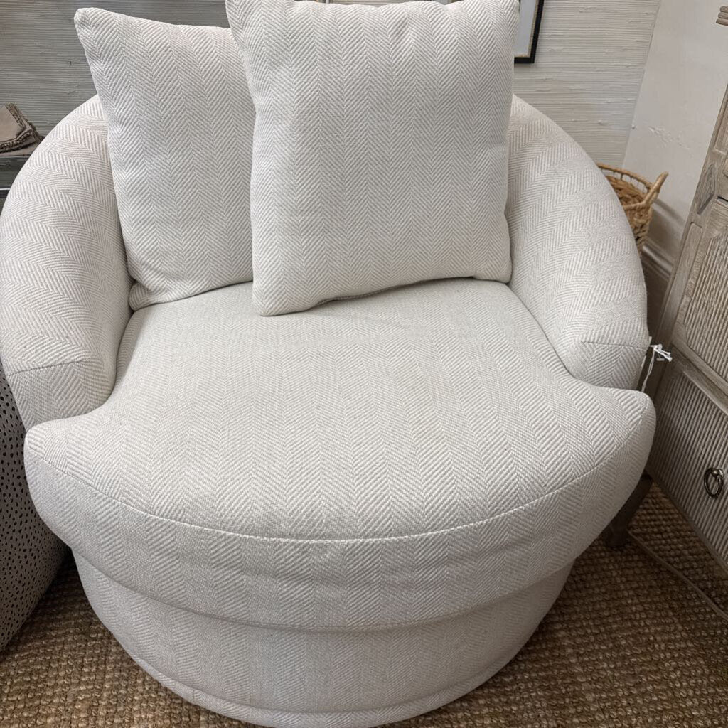 A round upholstered accent chair in a light beige herringbone fabric, featuring two matching square throw pillows, shown in a home interior setting.