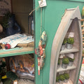A rustic boat-shaped wooden shelf display with small potted succulents, hanging from a teal-green wooden wall alongside a colorful fishing-themed ornament.