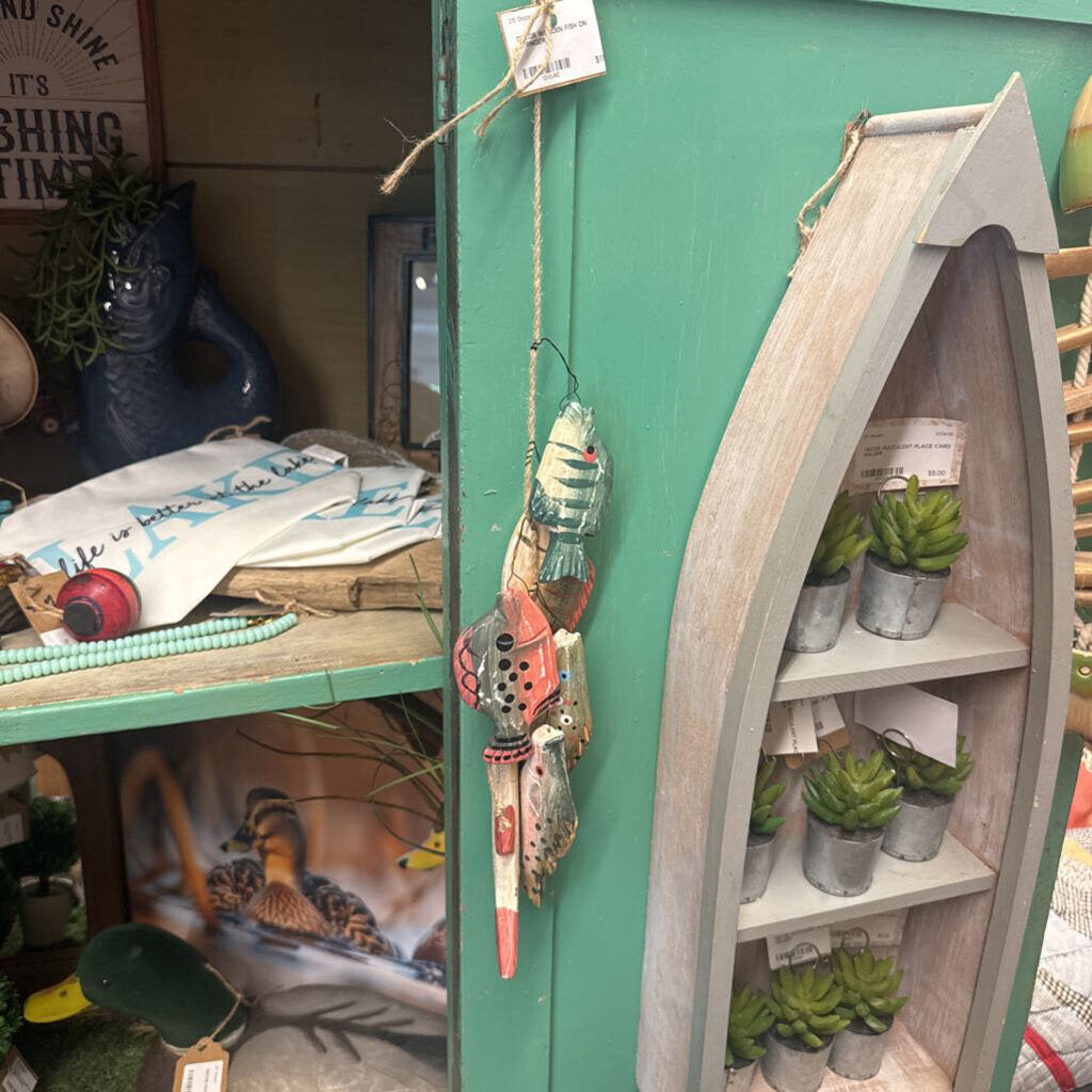 A rustic boat-shaped wooden shelf display with small potted succulents, hanging from a teal-green wooden wall alongside a colorful fishing-themed ornament.
