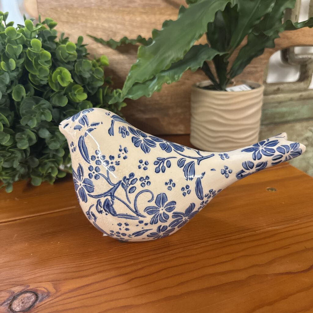 A ceramic bird figurine with a blue floral pattern on an off-white glaze, displayed on a wooden surface with green plants in the background.