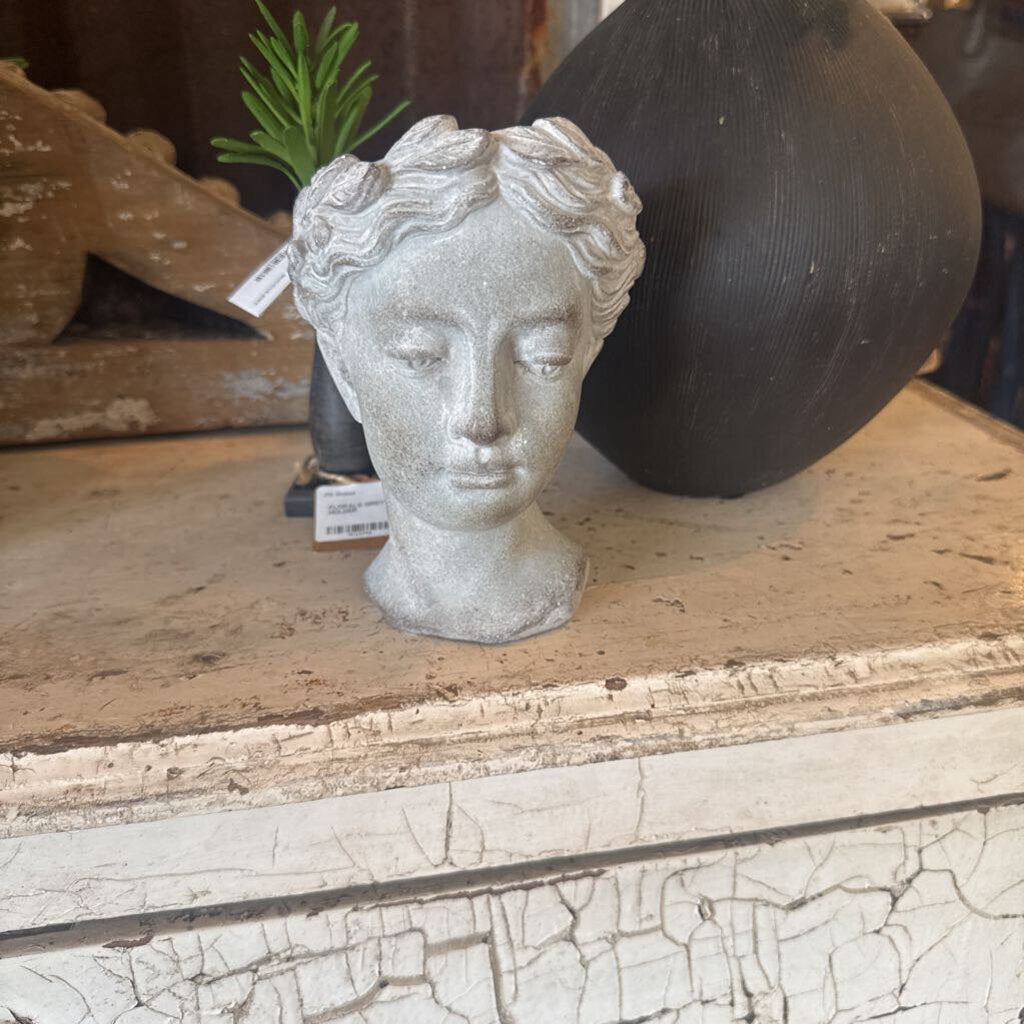 A small sculptural bust of a classical female head in a light gray stone or concrete finish, displayed on a distressed wooden surface next to a large black vase.