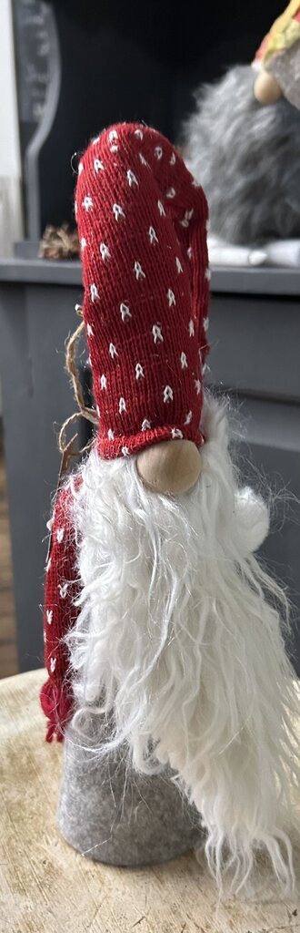 A decorative gnome figurine with a red knit hat and scarf patterned with small white motifs, a long white faux-fur beard, and a gray felt base, shown in a real indoor setting.